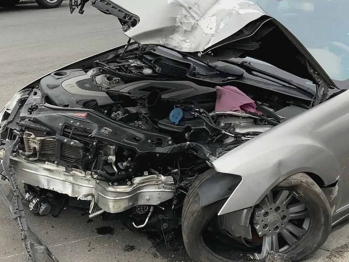 A silver sedan parked at a curb with its front end torn off, the hood crumpled, and the front wheel bent in